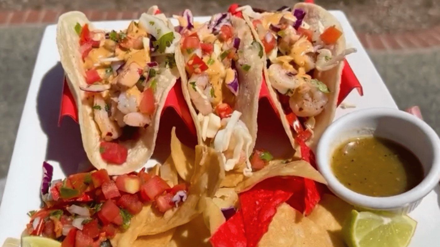 Three shrimp tacos with chipotle aioli, chips, pico de gallo and green salsa on a red plate.